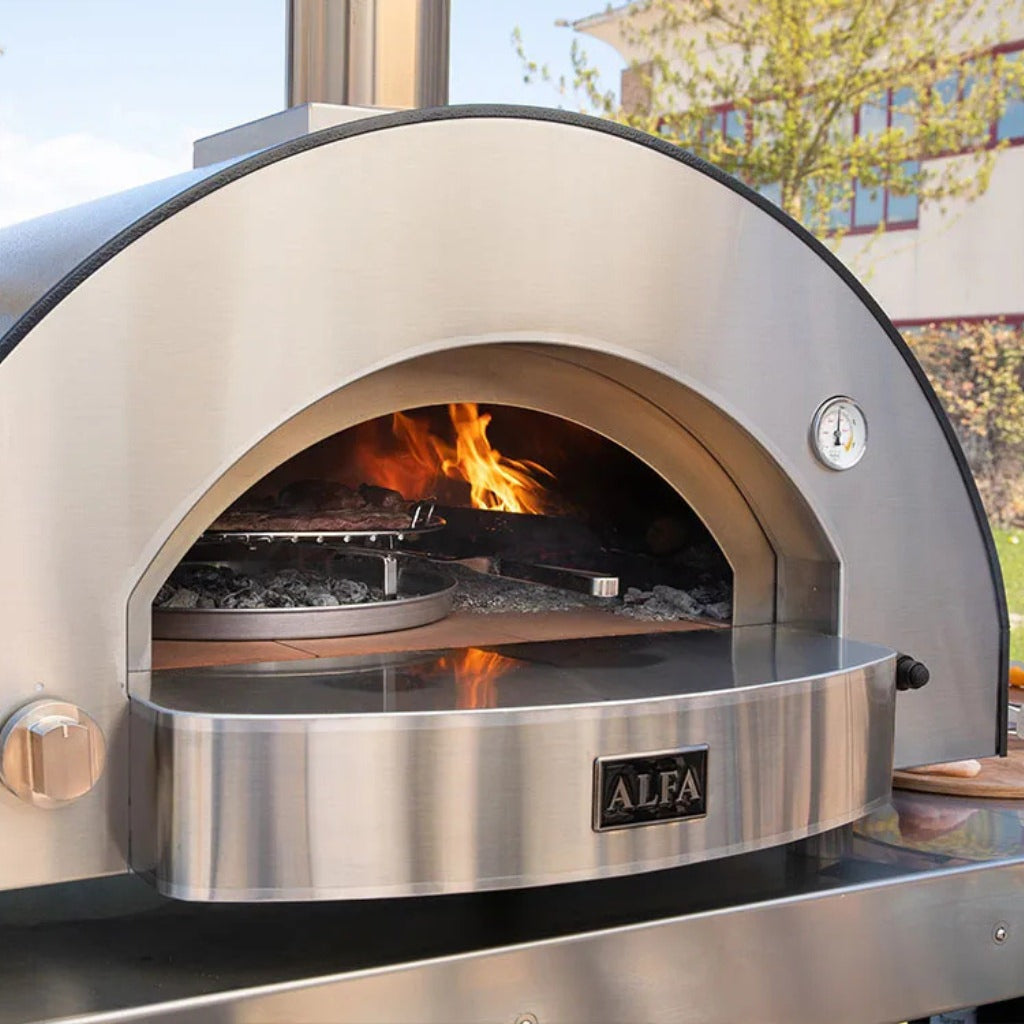 Alfa Forni BBQ 500 cooking meats in an Alfa stainless steel outdoor pizza oven with fire inside in an outdoor space.