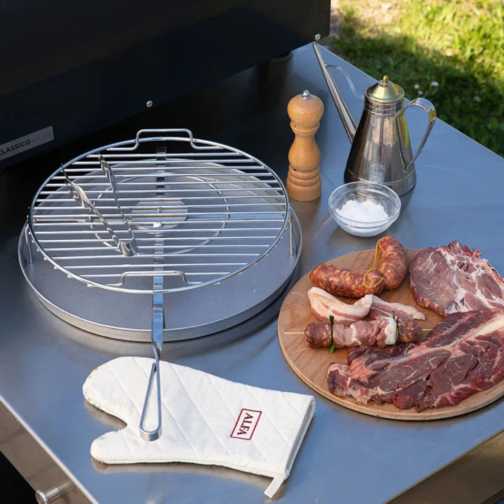 Alfa Forni BBQ grill with a wooden cutting board featuring raw meats and sausages, on a table outdoors.