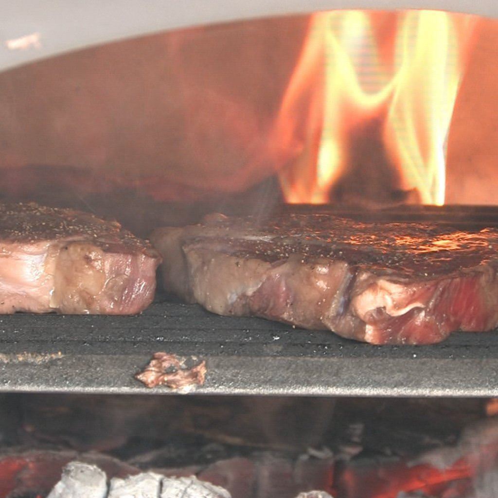 Chicago Brick Oven Hybrid Stand CBO 750 Freestanding Gas and Wood Fired Pizza Oven Cooking Steak with Fire in the Background of the Oven