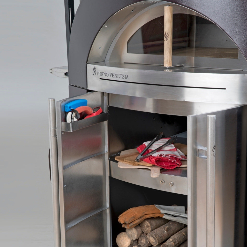 Forno Venetzia Bellagio 500 Mobile Wood Fired Pizza Oven in Copper with Doors Open on Mobile Cart Shelving with Cooking Accessories and Wood on Shelves.