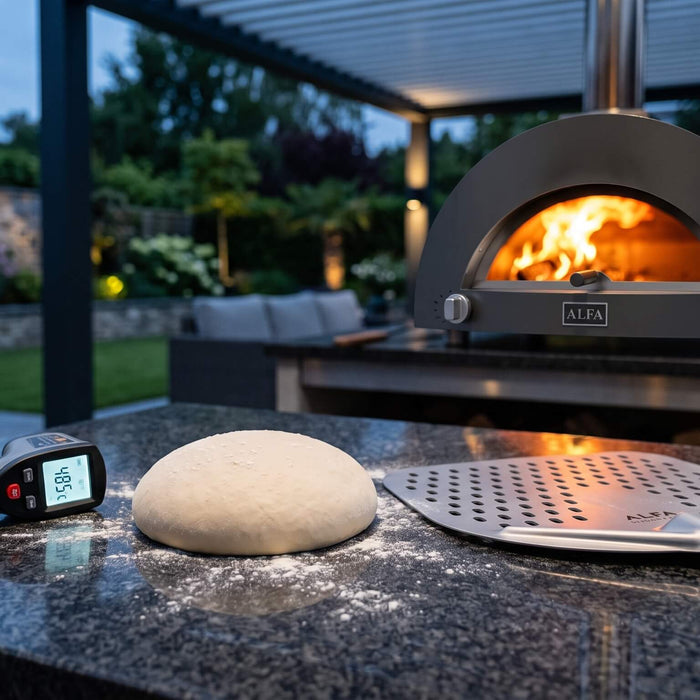 ALFA pizza oven with flames inside, with pizza dough, an infrared thermometer, and a perforated pizza peel on an outdoor kitchen counter.