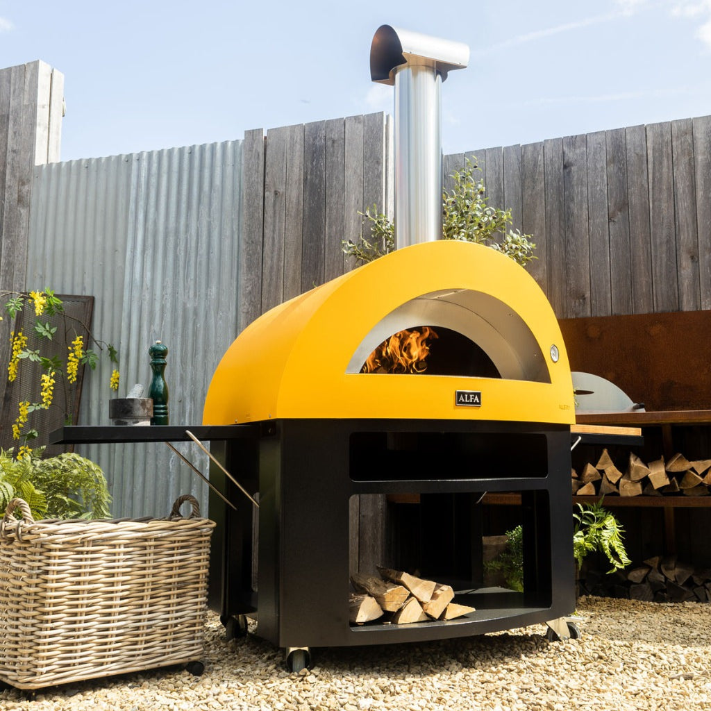 Alfa Allegro Pizza Oven Cart with yellow outdoor pizza oven on top with fire inside, set against a wooden fence and garden backdrop.