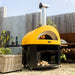 Alfa Allegro Pizza Oven Cart with yellow outdoor pizza oven on top with fire inside, set against a wooden fence and garden backdrop.