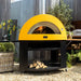 Alfa Allegro Pizza Oven Stand with yellow  outdoor pizza oven with fire inside, set against a rustic wooden fence and garden backdrop.