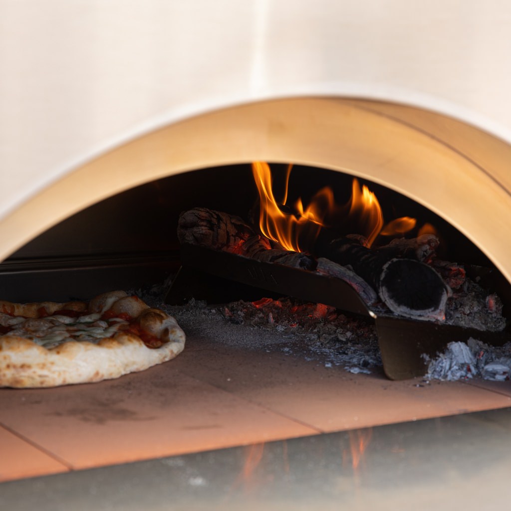 Alfa Forni Hybrid Kit inside wood-fired oven with pizza baking inside, flames visible on wood holder