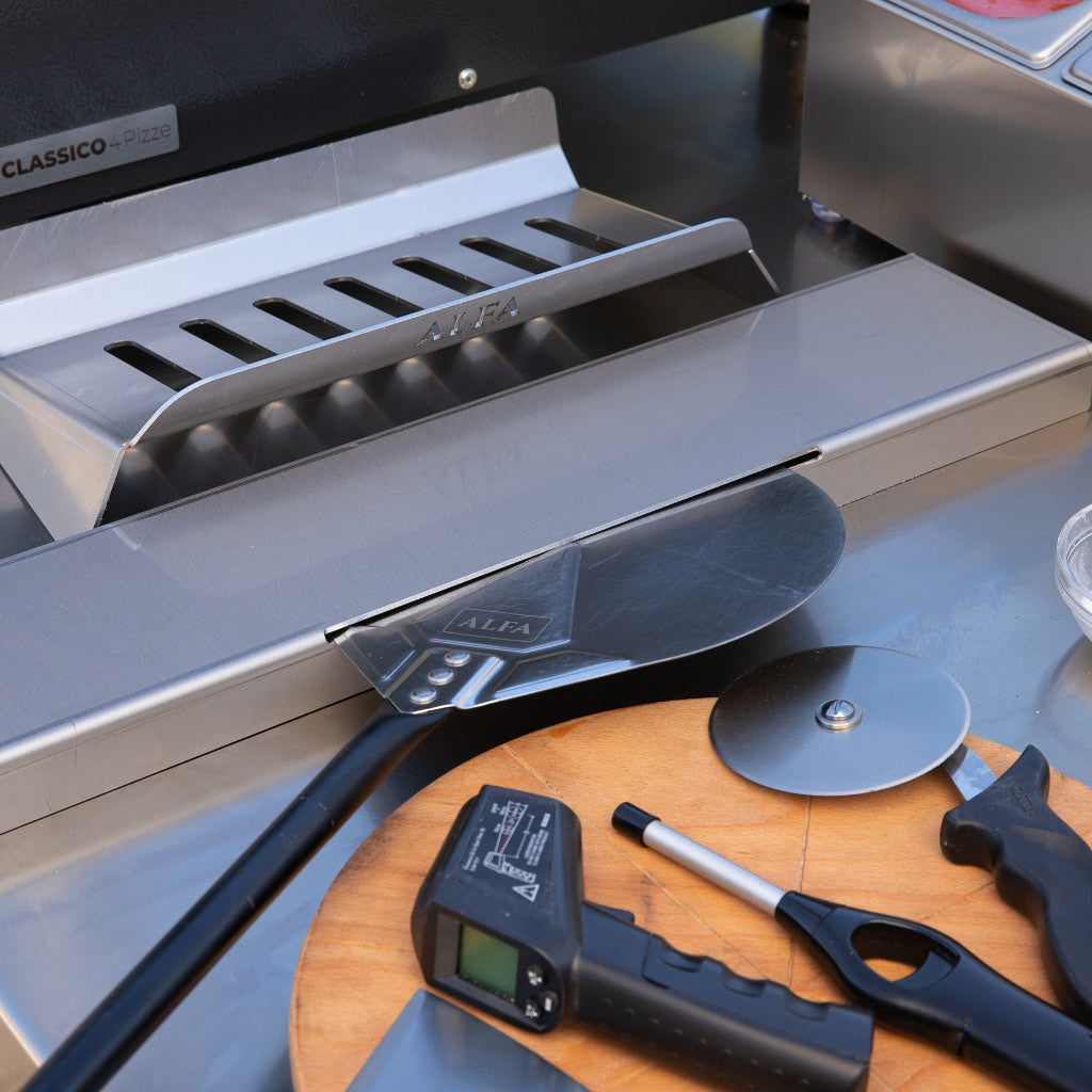 Alfa Forni Hybrid Kit close-up view on prep table with pizza cutter, digital thermometer, pizza peel, and a wooden cutting board with a pizza oven in the background.
