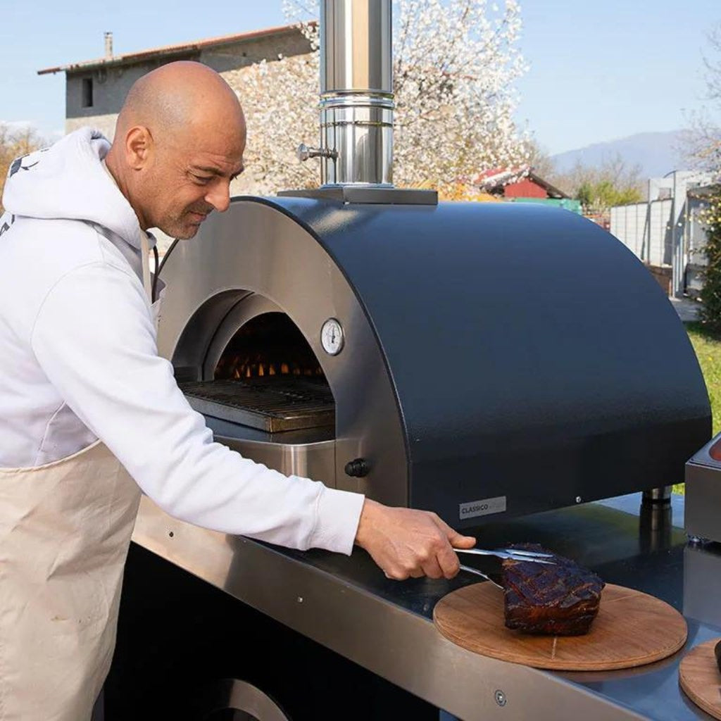 Alfa Forni Low and Slow Kit cooking ribeyes in an outdoor kitchen with a man placing a cooked ribeye on the table next to the oven with a beautiful outdoor background.