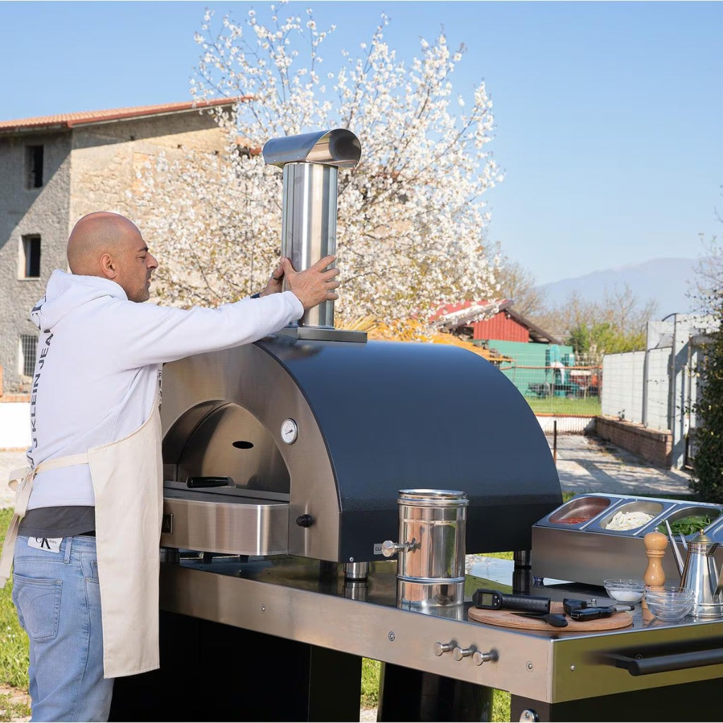 Alfa Forni Low and Slow Kit butterfly valve being installed by a man who is removing the existing flue to put it on the oven whil being outdoors with a scenic background.