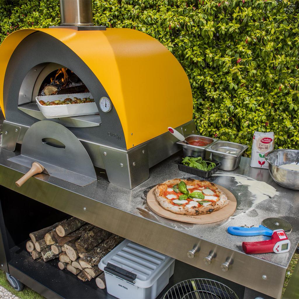 Alfa Forni Table with an outdoor pizza oven on top with a Margherita pizza on a wooden board, surrounded by ingredients and tools.