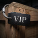 A black rectangular metal tag with silver 'VIP' lettering, attached to a silver cable lock, shown hanging on a rustic wooden pallet.
