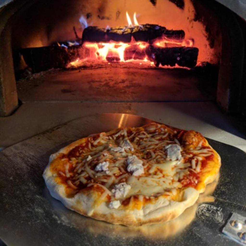 Chicago Brick Oven CBO 1000 Commercial Wood Fired Pizza Oven Kit Cooking Pizza with Wood Burning in the Oven in the Background